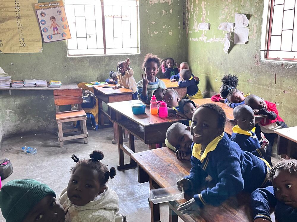 Viele Kinder in einem einfachen Klassenzimmer, einige lächeln in die Kamera, während ein älteres Kind ein Baby hält