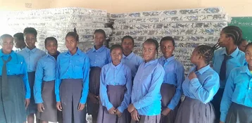 Eine Gruppe von Schülern in blauen Uniformen steht vor gestapelten Matratzen in einem Klassenzimmer