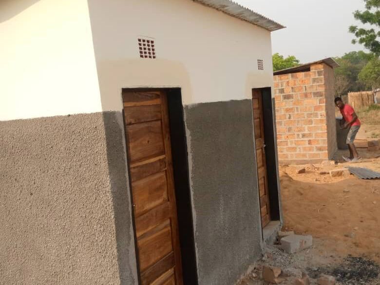 Neu gebautes Toilettengebäude mit zwei Holztüren und Wellblechdach, im Hintergrund ein Mann bei einem weiteren Rohbau