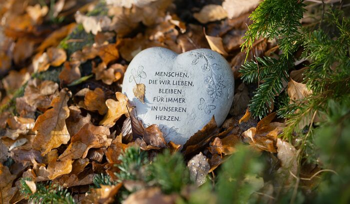 Herzförmiger Gedenkstein mit der Inschrift 'Menschen, die wir lieben, bleiben für immer in unseren Herzen', umgeben von Herbstlaub und Tannenzweigen