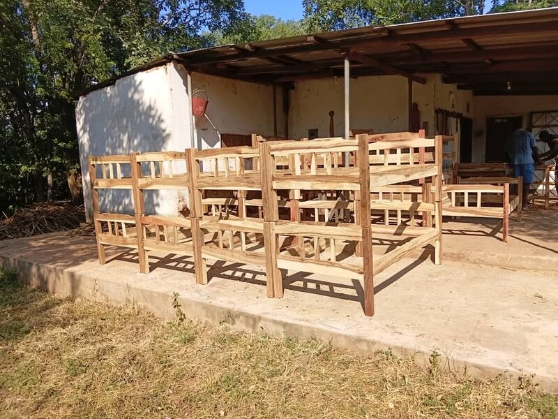 Mehrere neue, unlackierte Holz-Etagenbetten stehen vor einem einfachen Gebäude, im Hintergrund arbeiten Personen