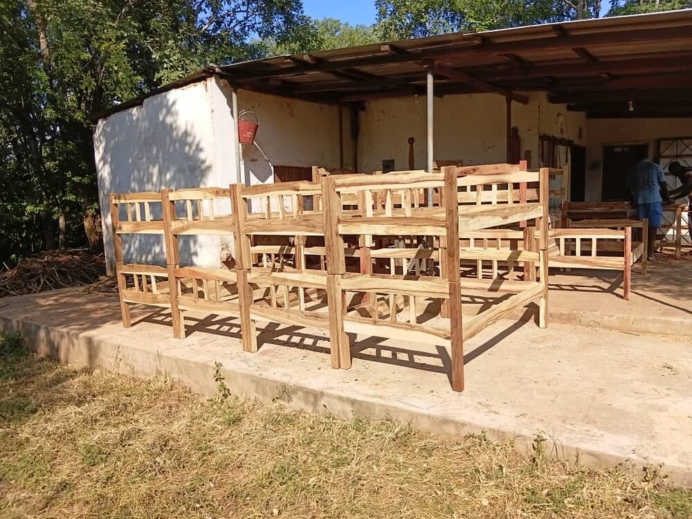 Mehrere neue, unlackierte Holz-Etagenbetten stehen vor einem einfachen Gebäude, im Hintergrund arbeiten Personen
