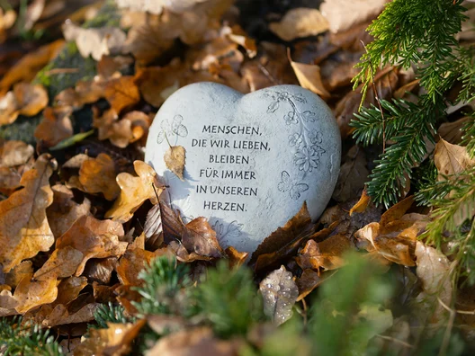 Herzförmiger Gedenkstein mit der Inschrift 'Menschen, die wir lieben, bleiben für immer in unseren Herzen', umgeben von Herbstlaub und Tannenzweigen