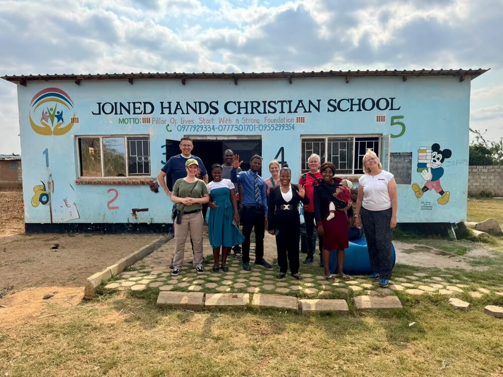 Eine Gruppe von Erwachsenen und einem Baby steht lächelnd und winkend vor der blauen "Joined Hands Christian School"