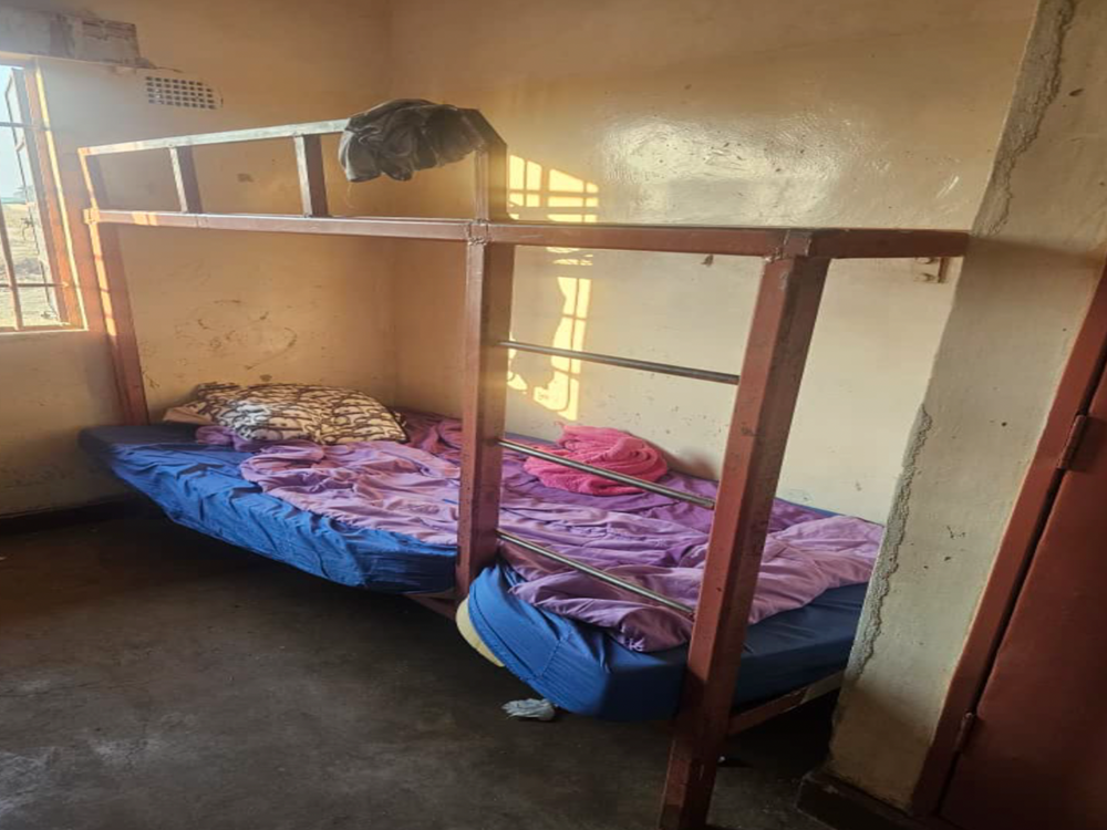 Ein einfaches Zimmer mit einem Metall-Etagenbett, dessen untere Koje mit blauer Matratze und lila Bettzeug ausgestattet ist, Sonnenlicht fällt durch ein Fenster
