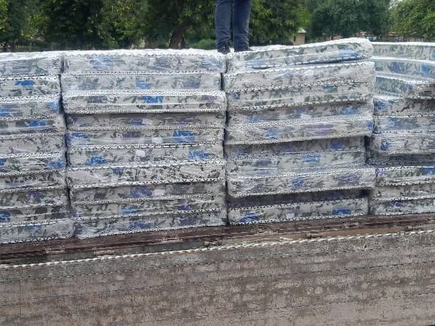Viele in Plastik verpackte Matratzen mit floralem Muster sind hoch gestapelt, die Beine einer Person sind oben sichtbar