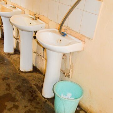 Several sinks in poor condition in a public washroom, with a bucket next to them.