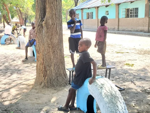 Mehrere Kinder spielen auf einem staubigen Platz mit bemalten Reifen und Bänken, im Hintergrund ein Gebäude und Bäume