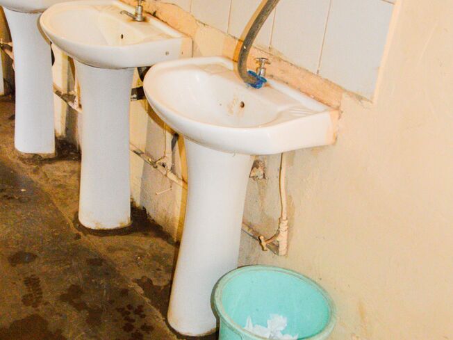 Mehrere Waschbecken in einem schlechten Zustand in einem öffentlichen Waschraum, mit einem Eimer daneben.