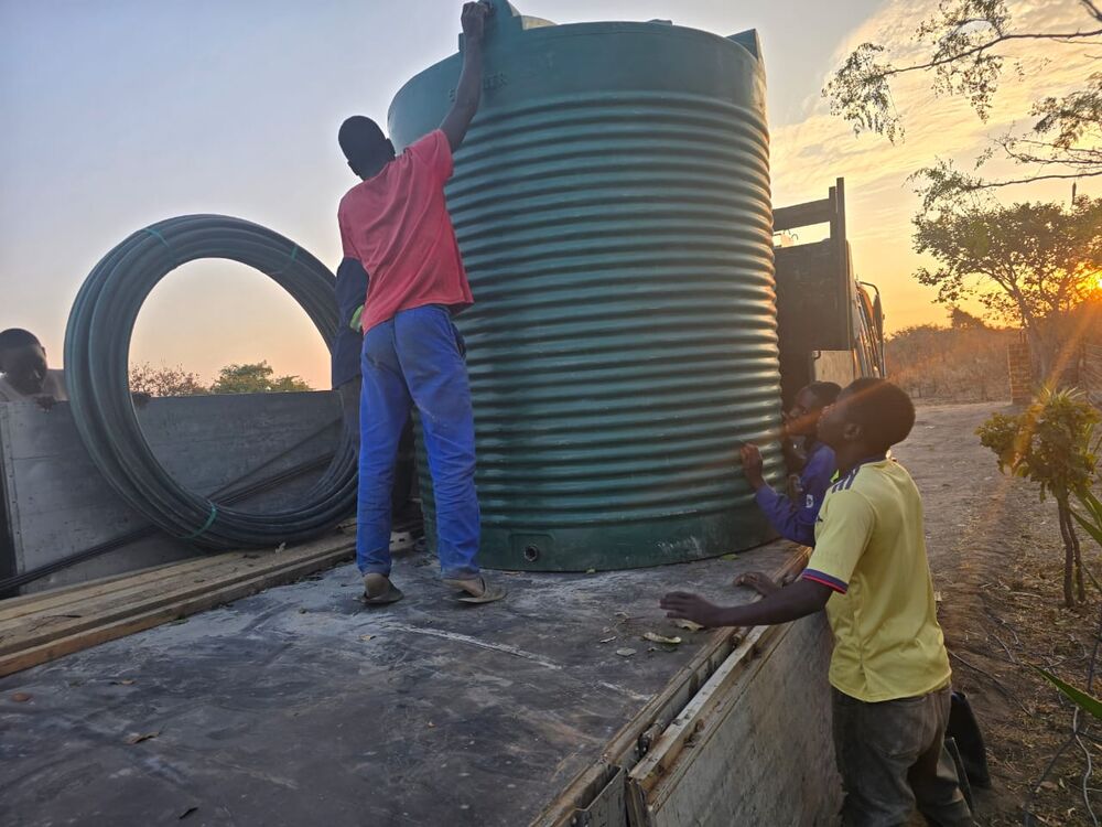 Mehrere Personen laden einen großen grünen Wassertank und schwarze Rohre auf die Ladefläche eines Lastwagens bei Sonnenuntergang