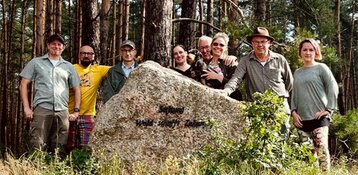 8 people are standing around a large stone in the forest and looking into the camera. It shows the club's team.