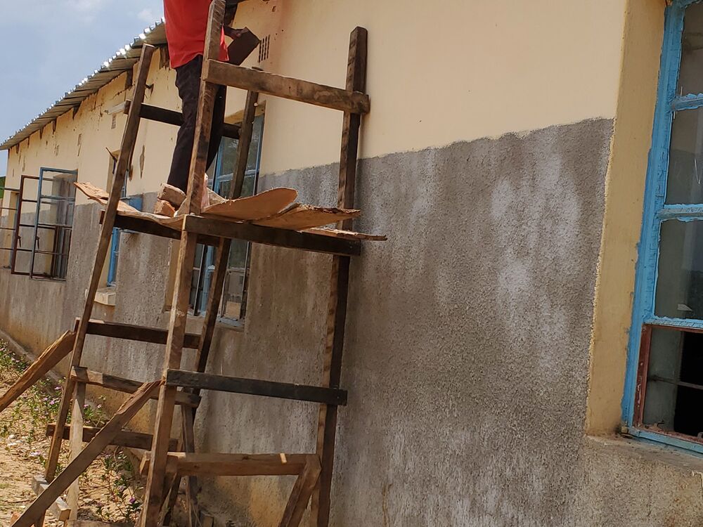 Ein Mann auf einem Holzgerüst arbeitet an der Dachtraufe eines Gebäudes mit hellgelber Wand und Wellblechdach