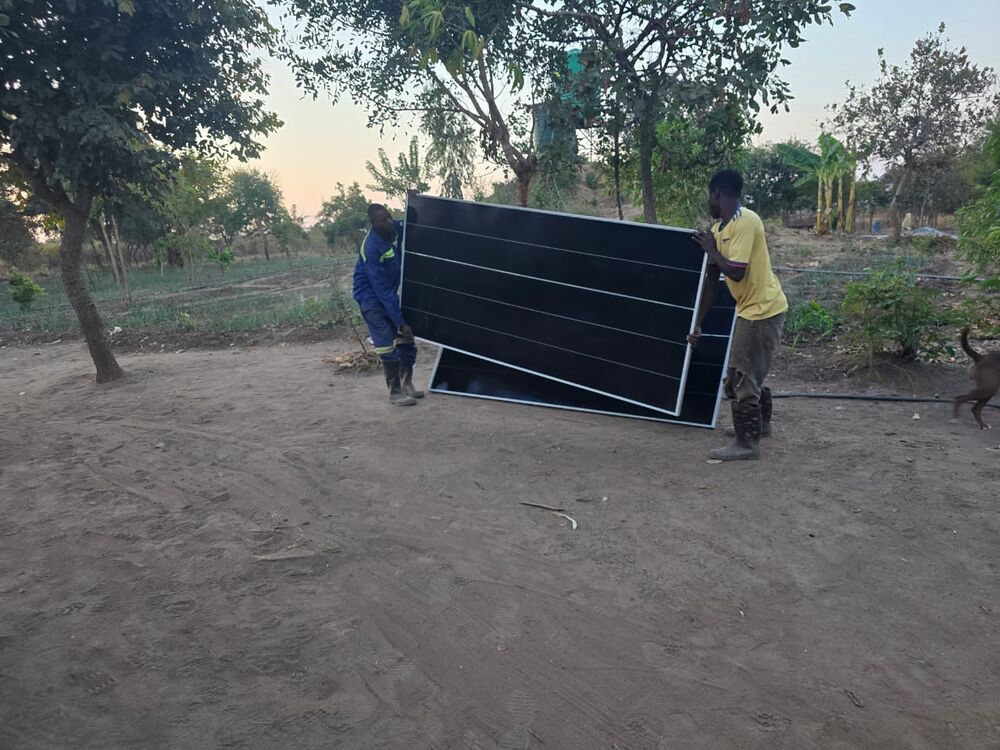 Zwei Männer tragen gemeinsam große schwarze Solarmodule über einen sandigen Boden in einer ländlichen Umgebung