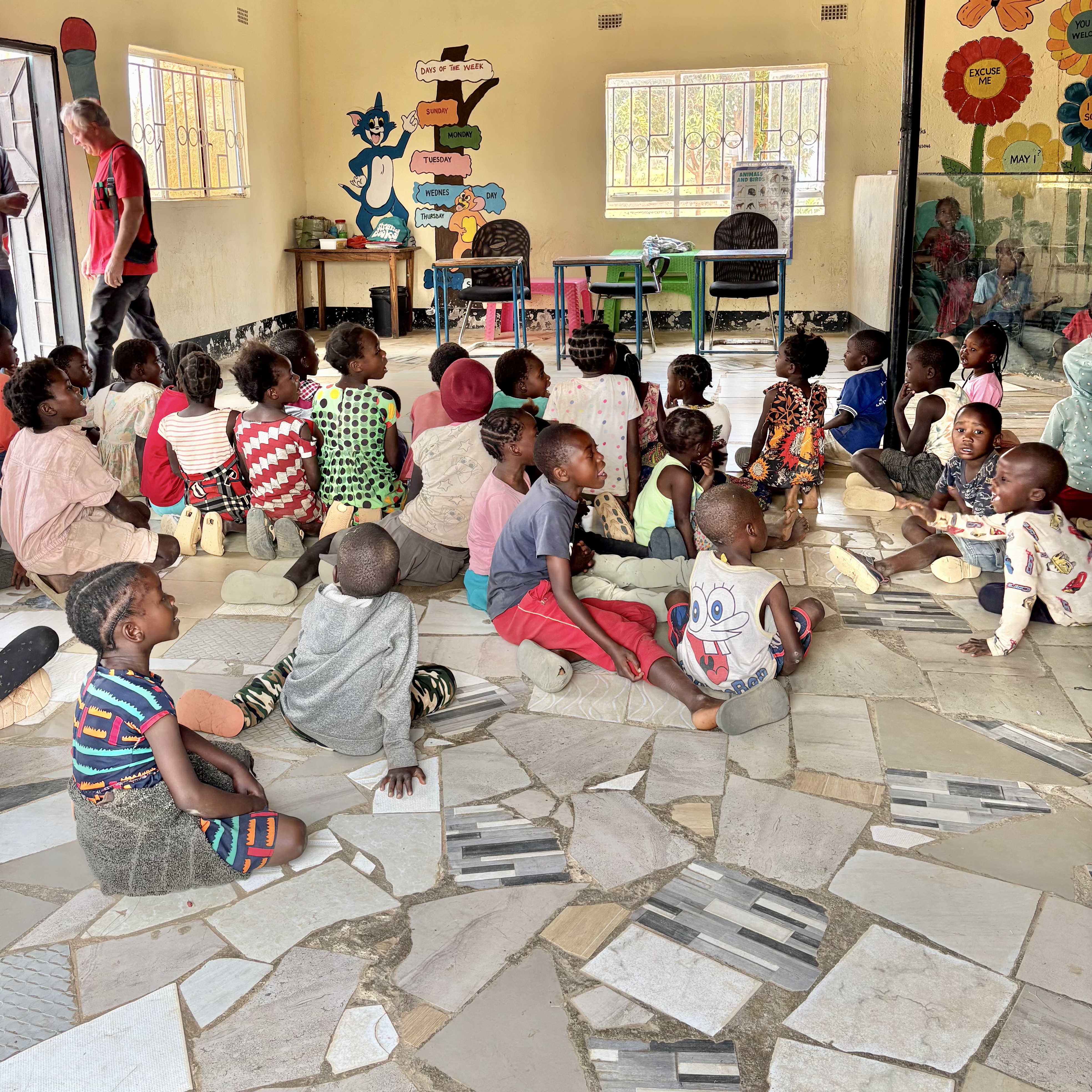 Viele Kinder sitzen auf dem Boden eines Klassenzimmers mit bunten Wandmalereien und blicken nach vorne